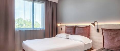 A modern hotel room with a large bed, a bedside table, and a view of the outdoors through a window.