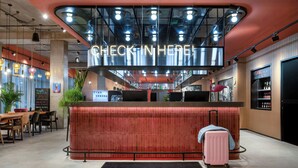 A hotel reception area with a 'Check-In Here' sign, a reception desk, a 'Nice to See You' mat, and a pink suitcase. - Moxy Verona (Verona)