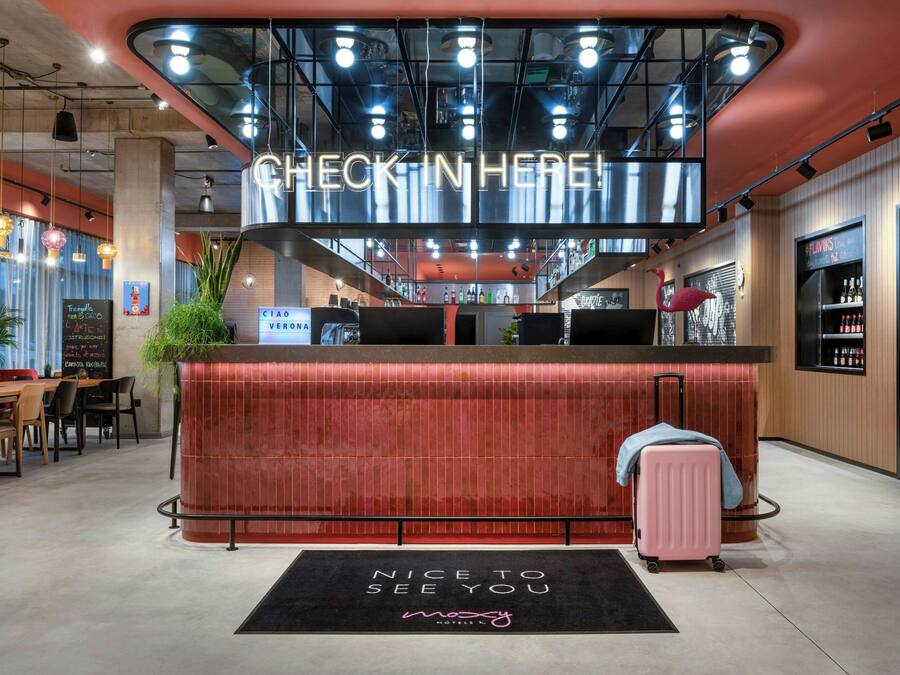Een hotel receptie met een bord waarop 'Check-In Here' staat, een receptiebalie, een vloermat met de tekst 'Nice to See You' en een roze koffer.
