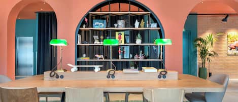 Un intérieur moderne avec une longue table en bois, quatre chaises, une bibliothèque contenant divers objets et deux lampes vertes.