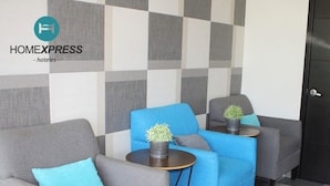 Een hotel lobby met een geruite wand, twee grijze fauteuils, een blauwe bank en een kleine tafel met een plant.