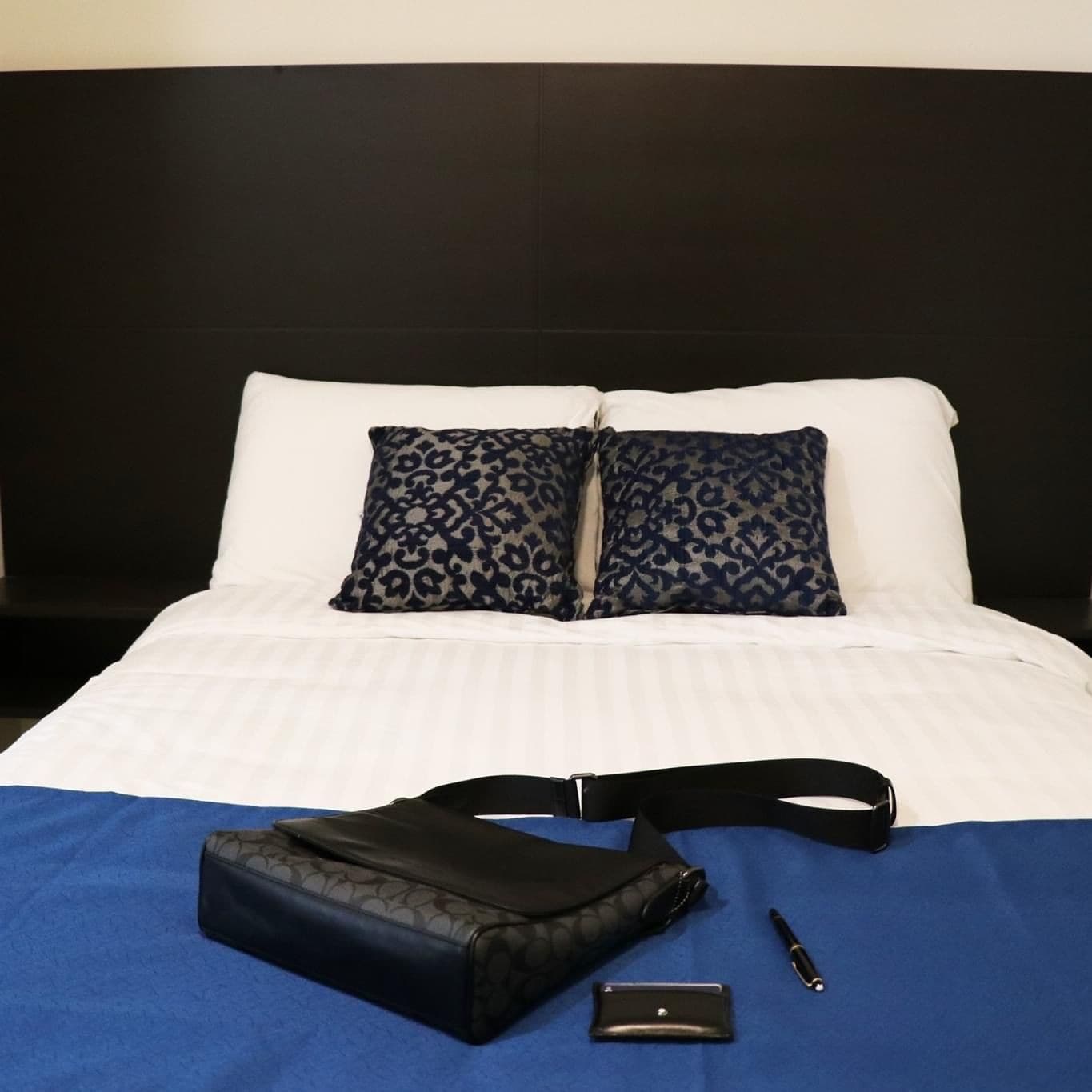 Una cama bien hecha con una almohada blanca y azul estampada, una bolsa negra, un bolígrafo y un objeto rectangular pequeño sobre una colcha azul.