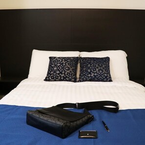 Una cama bien hecha con una almohada blanca y azul estampada, una bolsa negra, un bolígrafo y un objeto rectangular pequeño sobre una colcha azul.