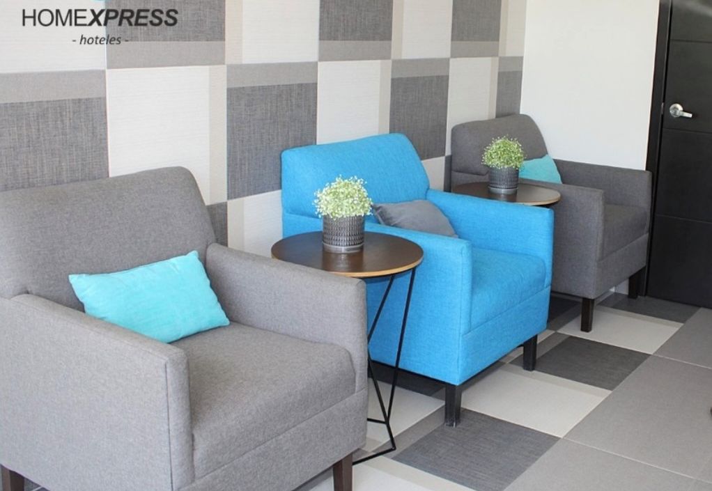 Un vestíbulo de hotel con una pared estampada de cuadros, dos sillones grises, un sofá azul y una pequeña mesa con una planta.