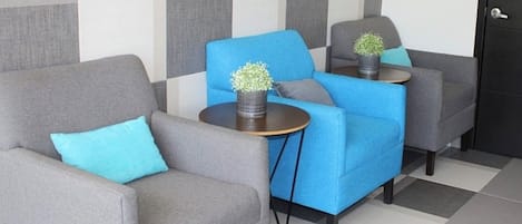 Un vestíbulo de hotel con una pared estampada de cuadros, dos sillones grises, un sofá azul y una pequeña mesa con una planta.