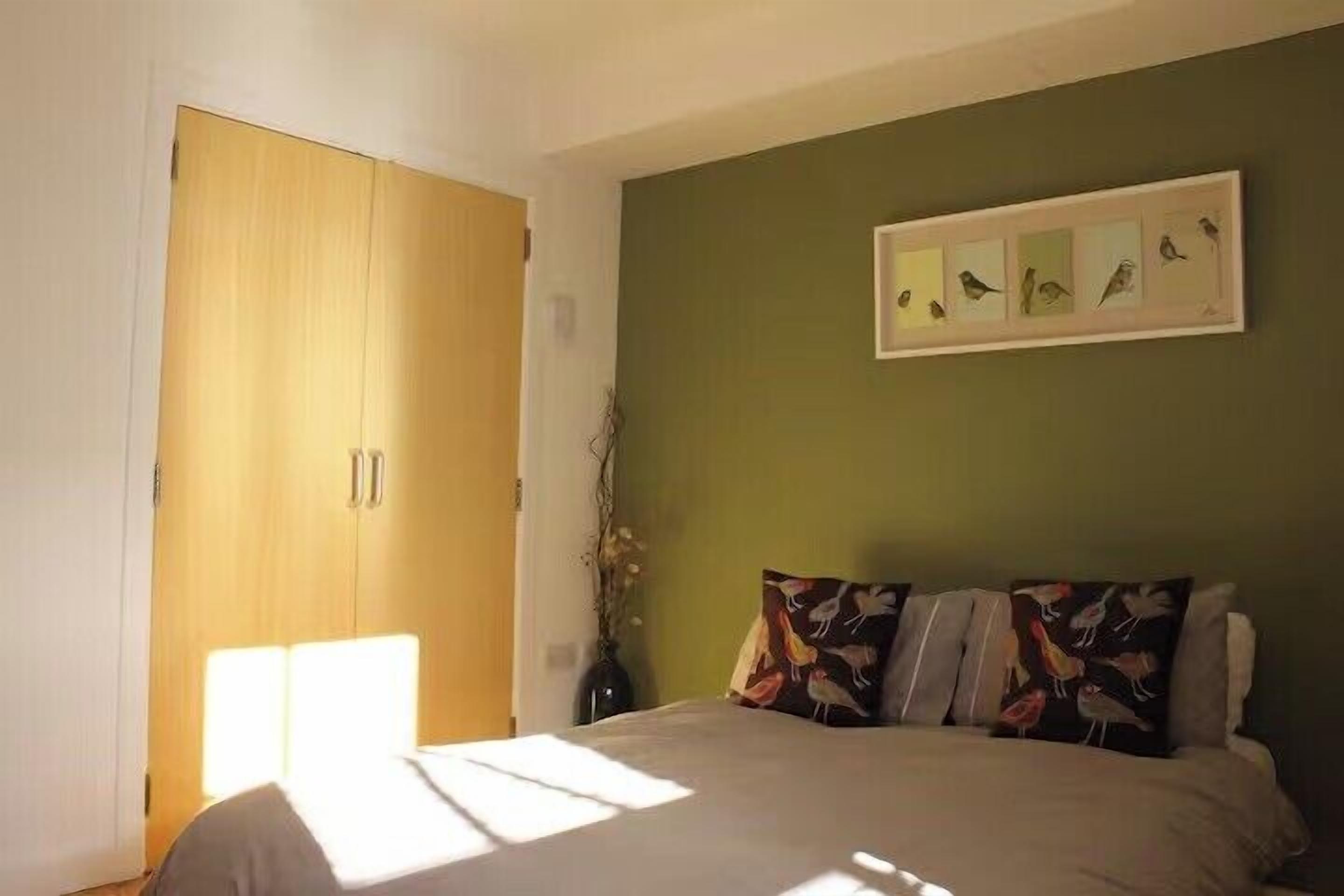 A bedroom with a bed, a green wall, a framed picture of birds, and a wooden wardrobe.