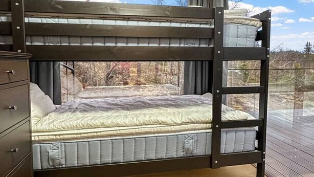 Una cama individual sobre otra con estructura de madera, un aparador y vistas a árboles a través de una ventana.