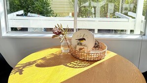 Une table ronde en bois recouverte d’une nappe jaune, un vase avec des fleurs séchées, une assiette décorative ornée d’un oiseau et un panier contenant une carte et une petite figurine.