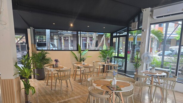 A modern cafe with wooden flooring, round tables, and chairs, surrounded by potted plants and large windows offering a view of the street. - h&Y chaweng (Koh Samui)