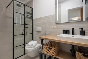 Un baño moderno con cabina de ducha de vidrio, inodoro blanco, tocador de madera con lavabo y espejo sobre el lavabo.