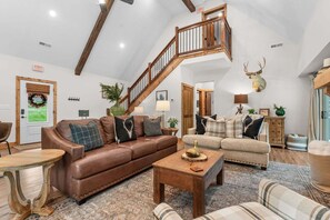 Un salón amplio con un sofá de cuero marrón, un sofá beige y una mesa de centro de madera. Una escalera de madera con barandilla conduce a otra habitación.