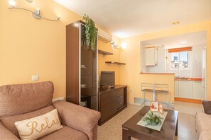 Een woonkamer met een bank, een salontafel, een televisie, een kast en een keukenhoek met kasten en een raam.