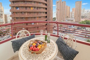 Un balcón con una mesa dispuesta con fruta y botellas, un mantel estampado y vistas a un paisaje urbano con edificios altos.