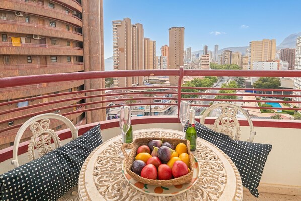 A balcony with a table set with a fruit basket, two wine glasses, and two bottles.