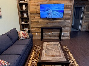 Ein Wohnzimmer mit einem dunkelbraunen Sofa, einem gemusterten Teppich, einer Holzwand mit einem darüber montierten Fernseher und einem Regal mit Dekorationen.