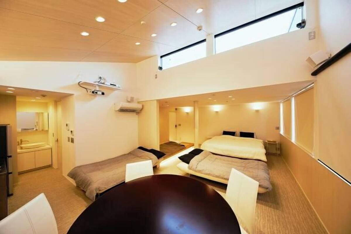 A compact hotel room with two single beds, a small table, a desk with a chair, a mirror, and a window providing natural light.