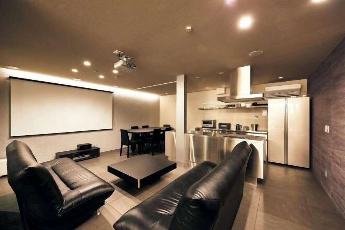 A modern living room with a large screen, leather sofas, a coffee table, and a kitchen area with a refrigerator and microwave.