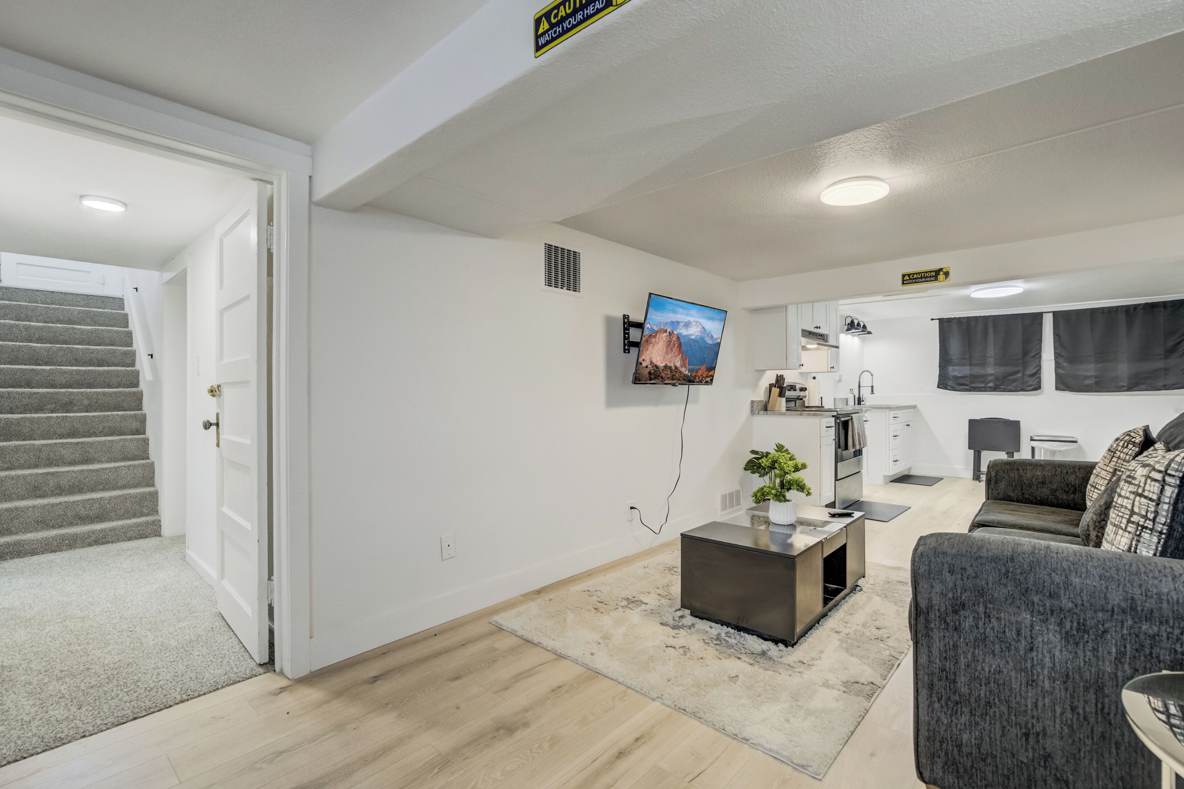 A modern living room with a staircase, a flat-screen TV, a kitchen area, and a grey sofa.