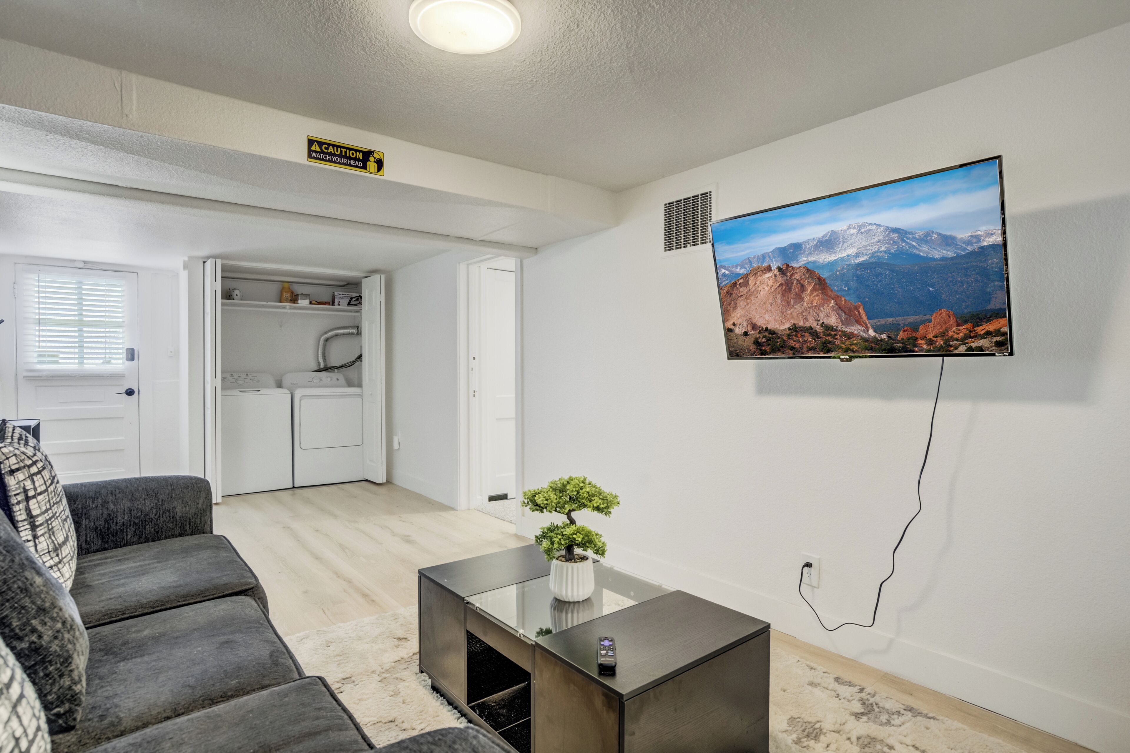 Una sala de estar moderna con un sofá, una mesa de centro con un bonsái, un televisor en la pared y una cocina al fondo.