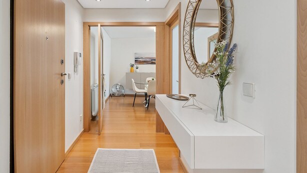 A modern hallway with wooden flooring, a white console table with a vase of flowers, and a large mirror with a geometric frame. - GuestReady - Modern apartment in Boavista (Boavista)