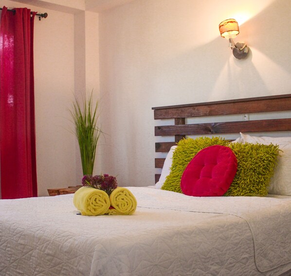 Un lit bien fait, recouvert d’une couette blanche, avec un oreiller rouge et un oreiller décoratif vert. Une lampe est fixée au mur au-dessus du lit, et une grande plante verte se trouve à côté.