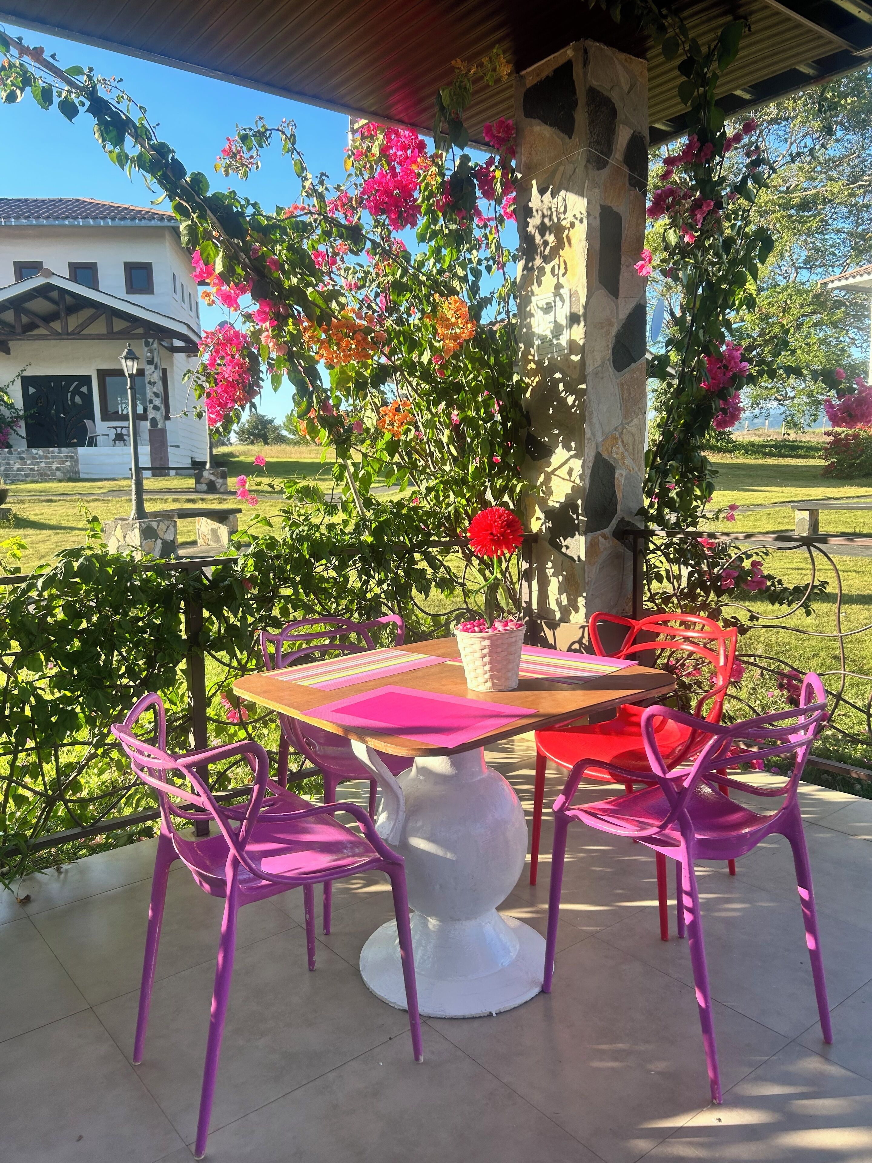 Un patio al aire libre con una mesa redonda y sillas moradas, rodeado de flores en flor y un pilar de piedra.