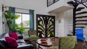 A modern living room with a black leather sofa, green patterned chairs, a round wooden table with a bowl of fruit, and a staircase with a black railing. - Country Hills Las Lajas (San Felix)