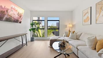 A modern living room with a glass coffee table, a sofa, a bench, and a large window offering a city view.