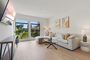 A modern living room with a white sofa, a glass coffee table, and a TV mounted on the wall.