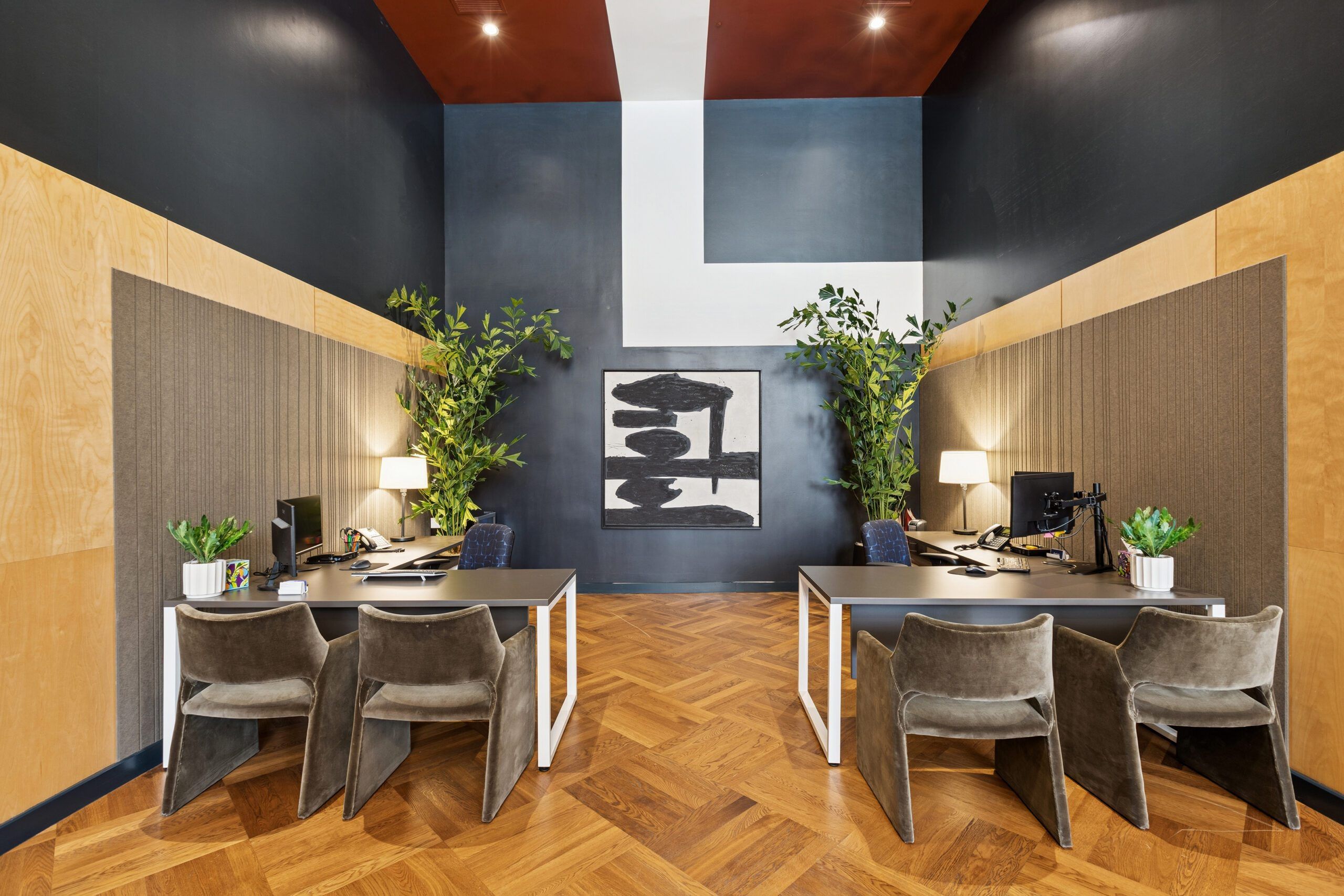 A conference room with a wooden herringbone floor, two desks with computers, chairs, and potted plants.