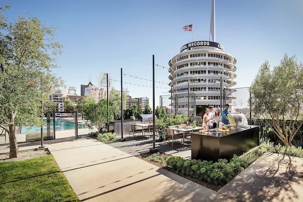 Un espace piscine aménagé sur le toit, avec un barbecue et des sièges, entouré de verdure et offrant une vue sur un immeuble portant l’inscription « CATHOL RECORDS ».