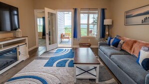 A living room with a grey sofa, a coffee table, a TV, and a large window with curtains. - Branson Fun Zone | Dog-Friendly • Games • Pool (Branson)