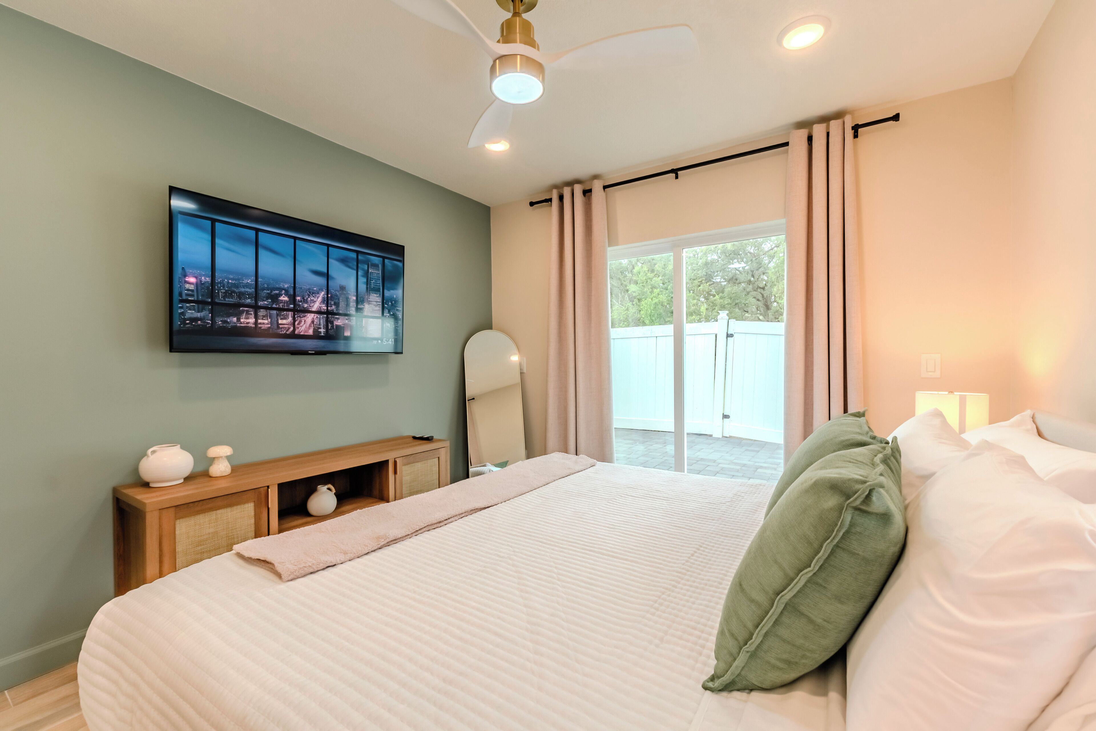 A bedroom with a large bed, a flat-screen TV mounted on the wall, a ceiling fan, and a sliding glass door leading to an outdoor area.