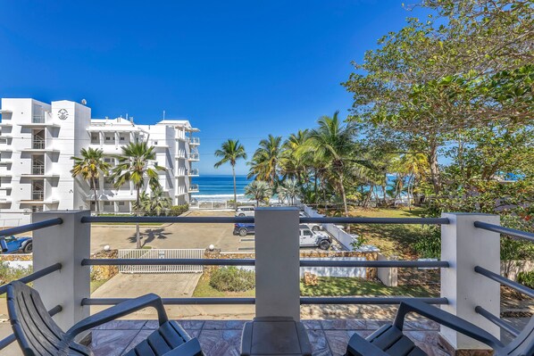 A balcony with a view of a beach and palm trees, overlooking a parking area and a white building with balconies.