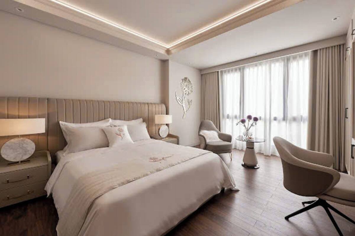 Habitación de hotel con una cama grande, dos mesitas de noche, una silla, una mesita con un florero y un ventanal con cortinas.