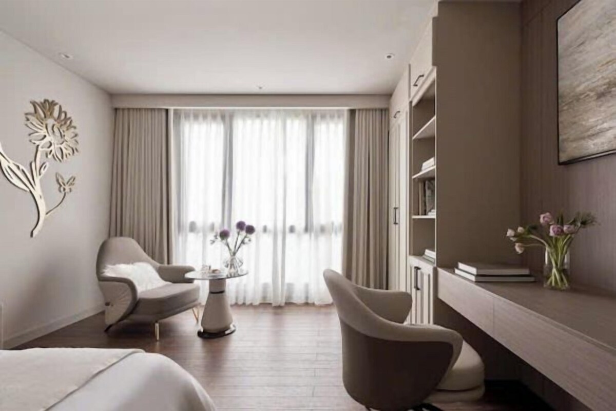 Habitación de hotel moderna con un ventanal grande, dos sillones, una mesita redonda y una estantería.