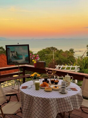 Ein Tisch gedeckt mit Frühstücksangeboten, ein Gemälde und Blick auf das Meer bei Sonnenuntergang.