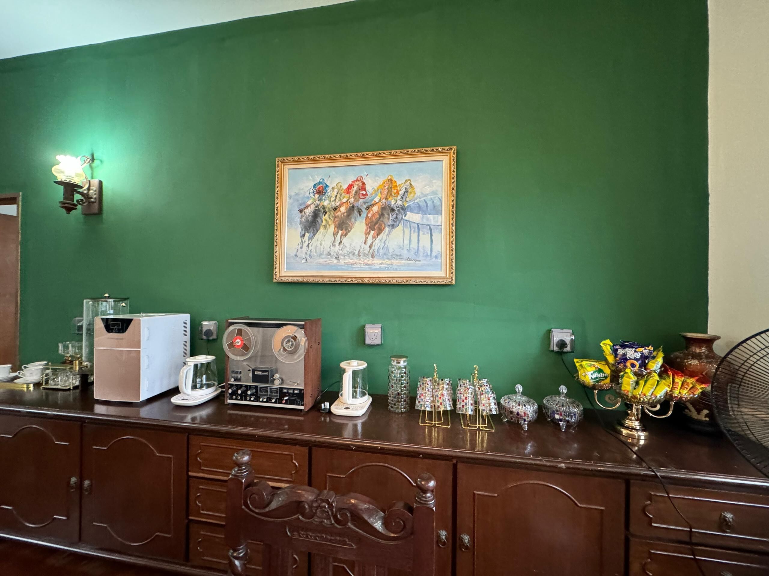A room with a green wall, a framed painting of a horse race, a vintage radio, a coffee machine, and decorative glassware.