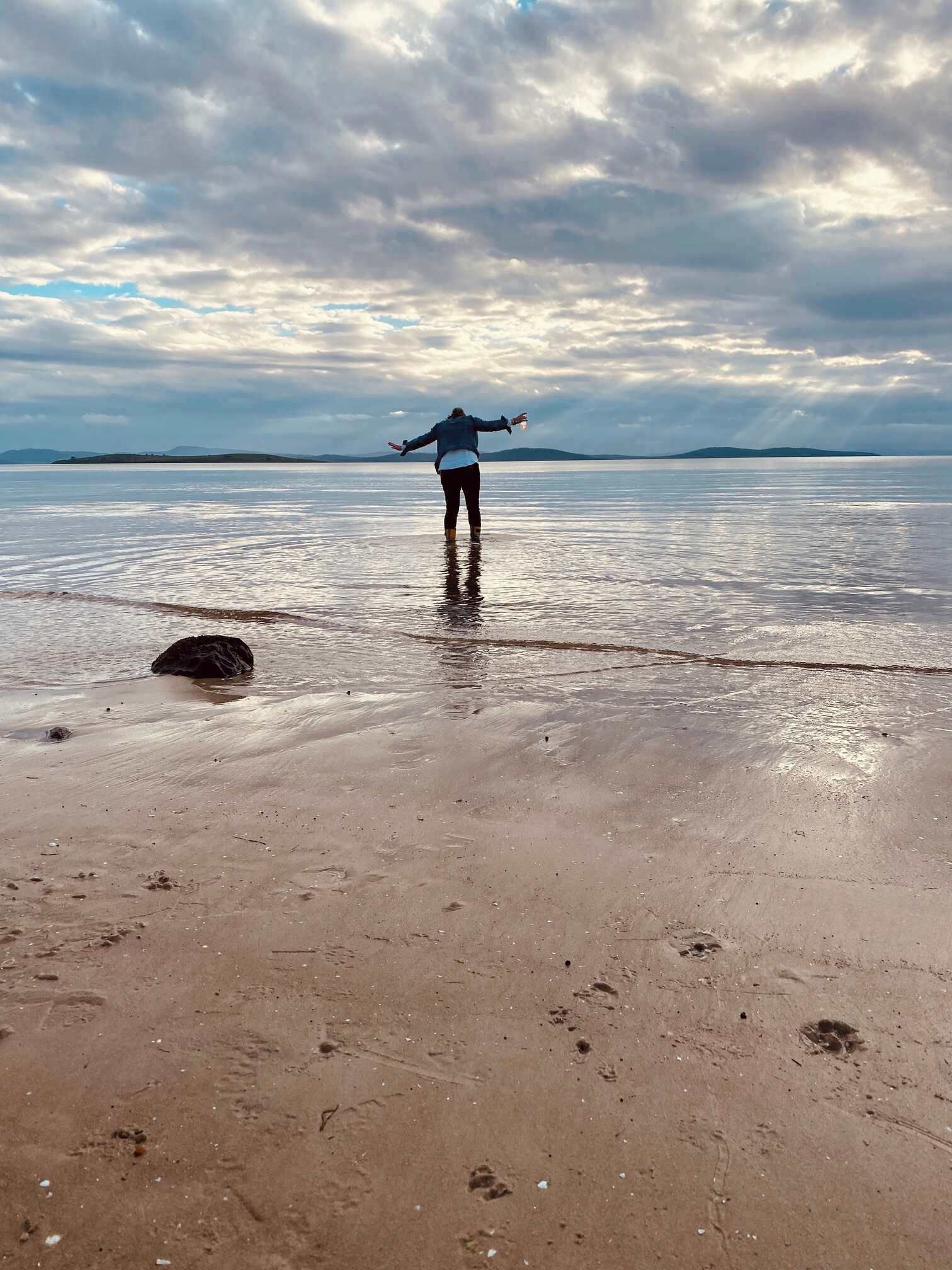 A person standing on a beach with a vast body of water and distant land in the background.