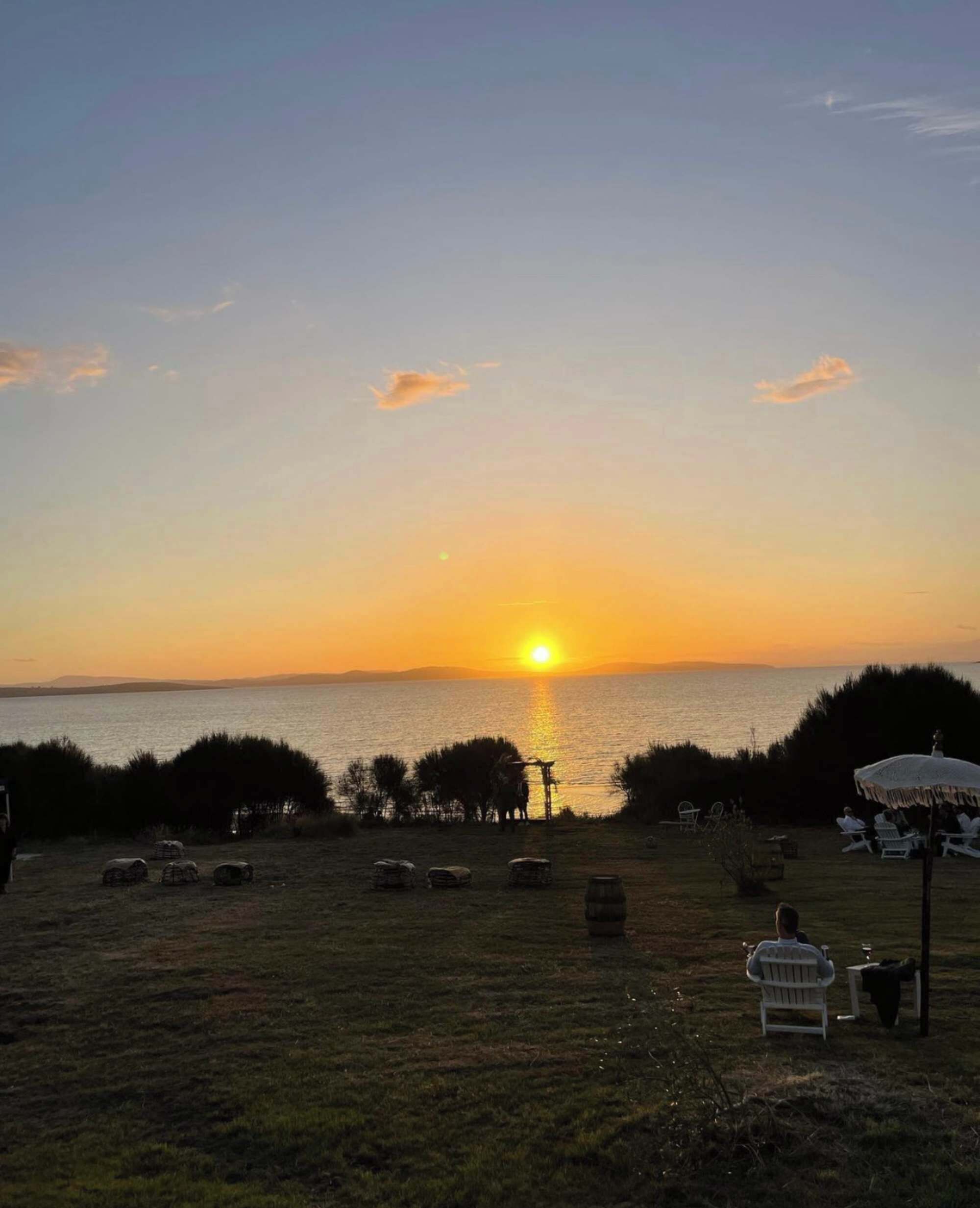 A sunset view over a body of water with a grassy area, some seating, and a few people.