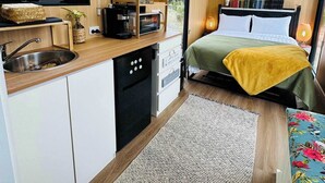 Une chambre compacte et moderne, dotée d’une kitchenette, d’un lit recouvert d’une couverture verte et d’une grande fenêtre donnant sur un paysage verdoyant.