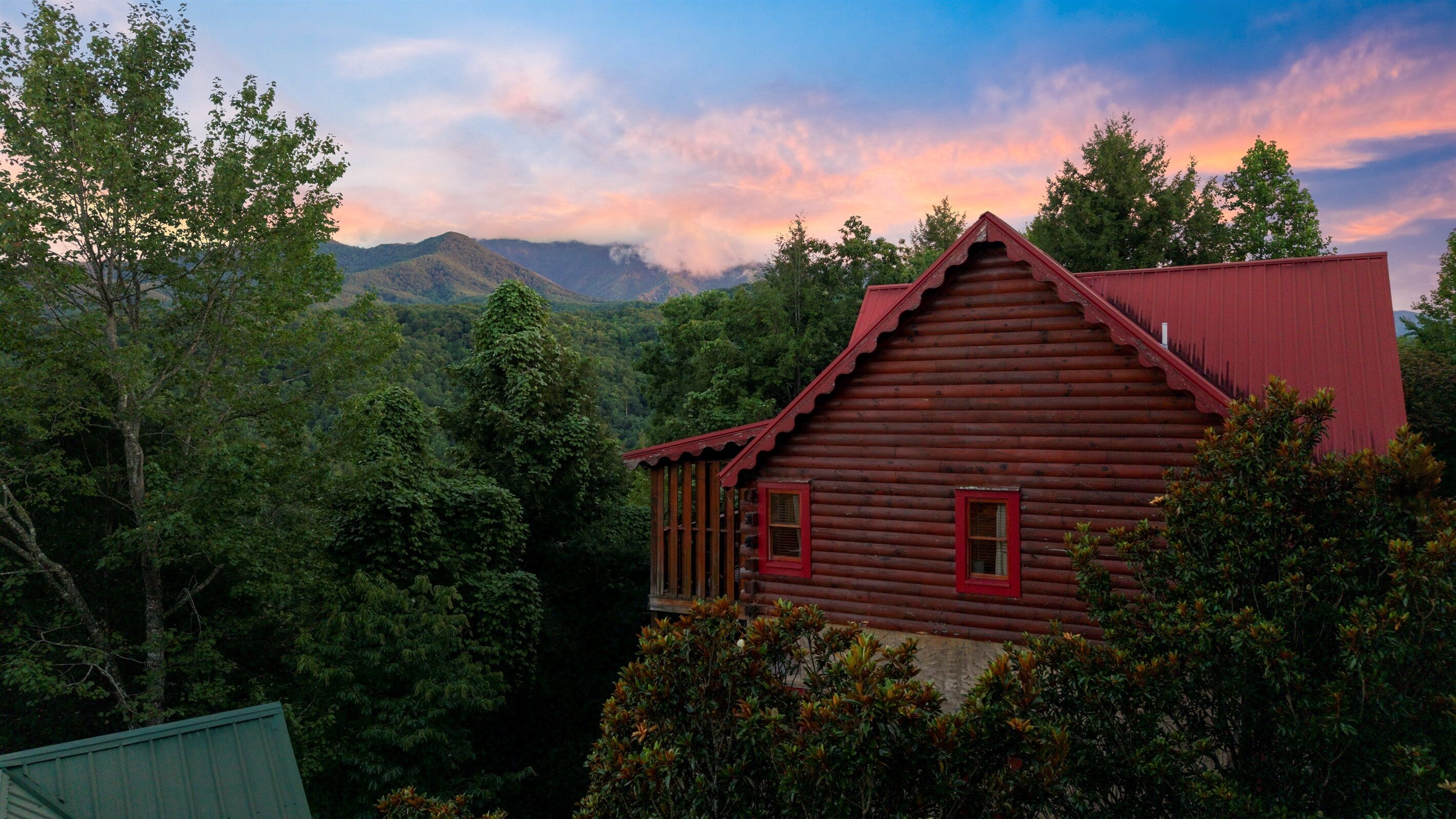 Une cabane en rondins avec un toit rouge, entourée d’une forêt dense et avec des montagnes en arrière-plan.