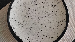 Une assiette blanche parsemée de petites taches noires, posée sur un tapis texturé, une tasse et une soucoupe blanches, et un verre transparent sur pied, le tout sur un plan de travail foncé.