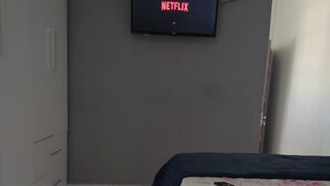 Une chambre à coucher avec un lit, un téléviseur fixé au mur qui affiche le logo « NETFLIX », et une télécommande posée sur le lit.