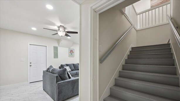 A modern living room with a grey sofa, a ceiling fan, and a staircase with a white railing.