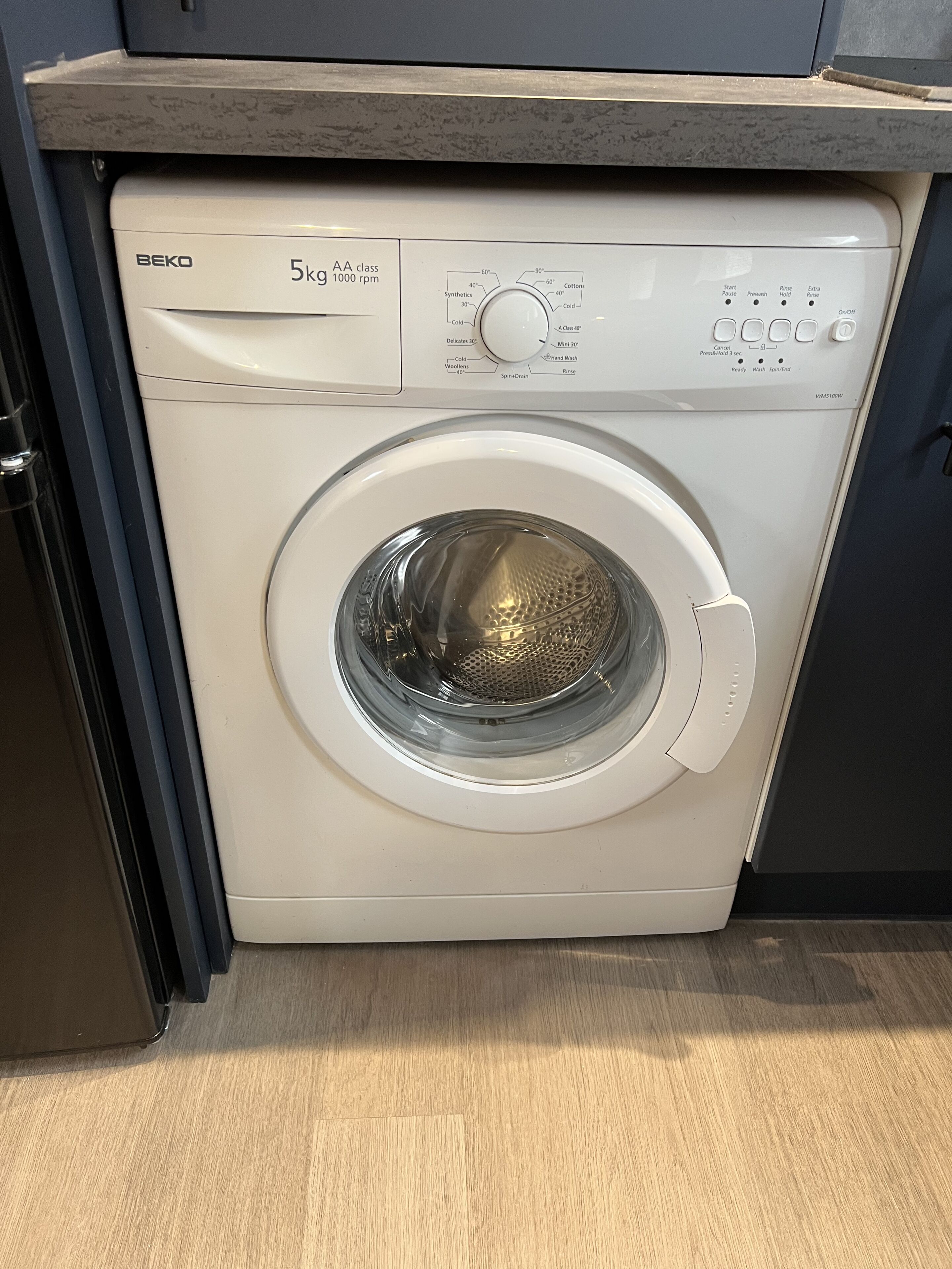 A front-loading BEKO 5kg washer with a control panel, located in a kitchen with a wooden countertop and a dark cabinet.