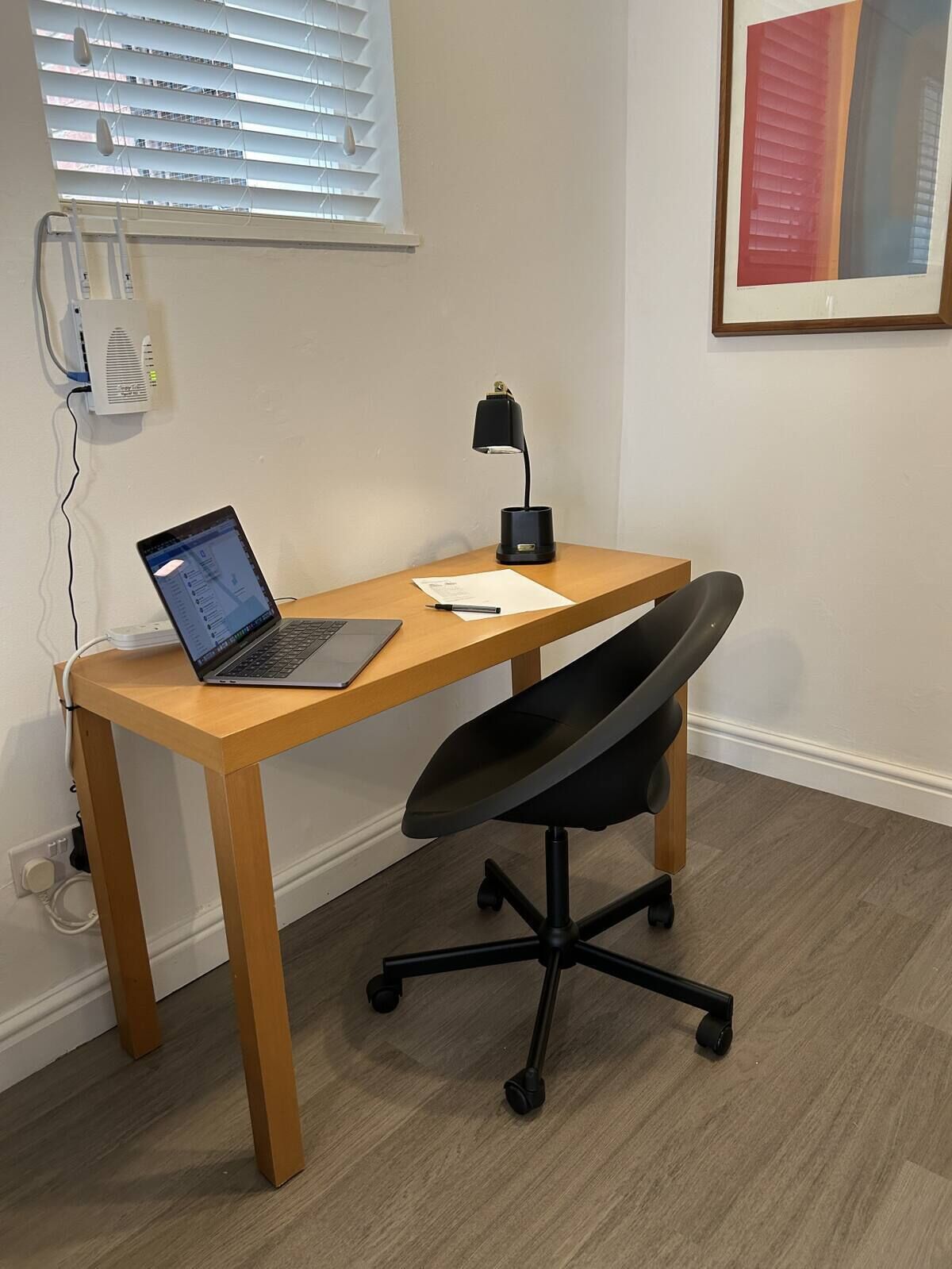 A wooden desk with a laptop, a black office chair, a desk lamp, and a framed picture on the wall.