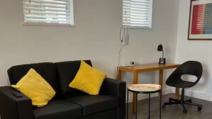 Ett modernt vardagsrum med en svart soffa, två gula kuddar, ett litet runt bord, en svart kontorsstol och ett skrivbord i trä med en lampa.