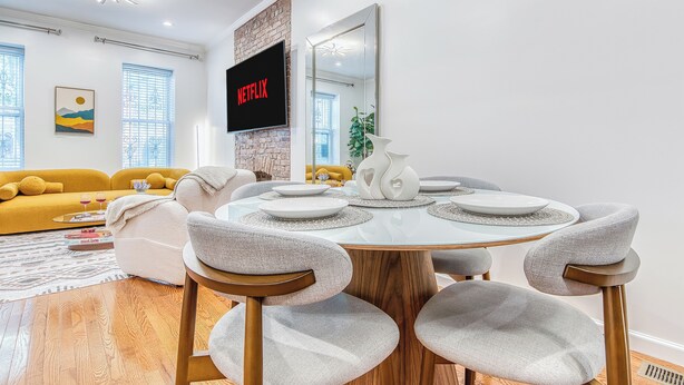 Una sala de estar moderna con una mesa de comedor redonda, sillas tapizadas en gris, un sofá amarillo y un televisor de pantalla plana montado en una pared de ladrillo.
