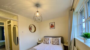 A neatly arranged bedroom with a bed, bedside tables, a hanging light fixture, a mirror, and a window with a plant. - Comfortable 2 Bedroom House in Bracknell (Bracknell)